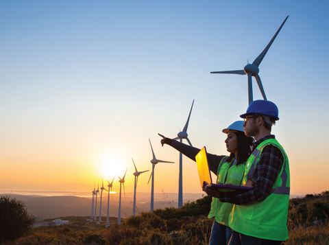 Two people wearing high vis and PPE stood in a wind turbine field holding a laptop and pointing to the distance.